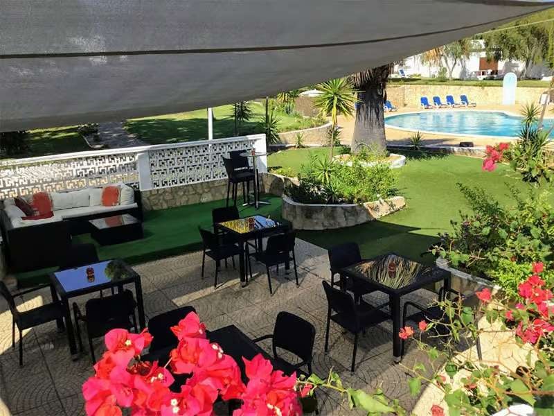 Outdoor poolside lounge area at Prado Vilamoura with shaded seating, bougainvillea flowers, and a swimming pool with sun loungers.