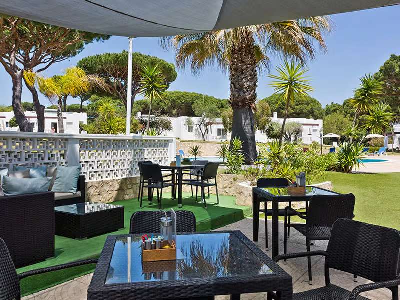 Poolside lounge area at Prado Vilamoura with wicker furniture and tropical plants, overlooking a pool and villas.