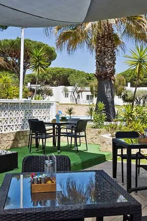 Poolside lounge area at Prado Vilamoura with wicker furniture and tropical plants, overlooking a pool and villas.