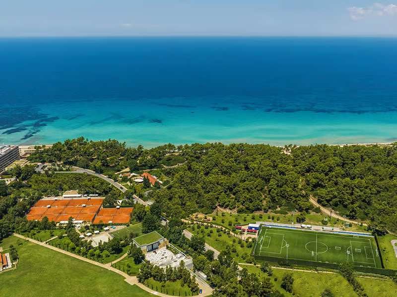 Aerial view of tennis courts at Sani Beach School, with greenery and the ocean in the background.