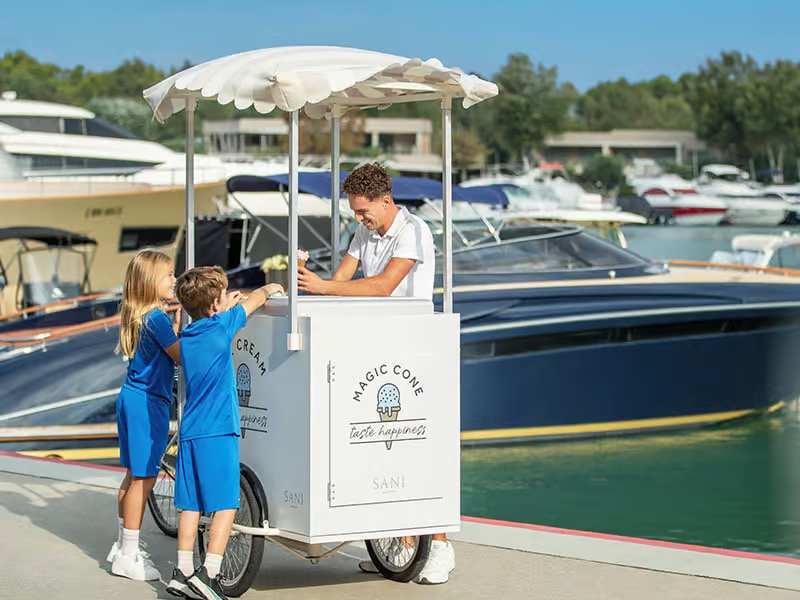 Children in school uniforms buying ice cream from a white cart marked 'Magic Cone' near yachts at a marina.