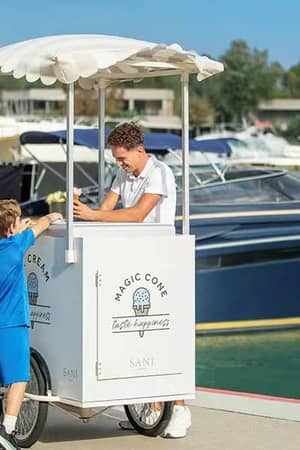 Children in school uniforms buying ice cream from a white cart marked 'Magic Cone' near yachts at a marina.