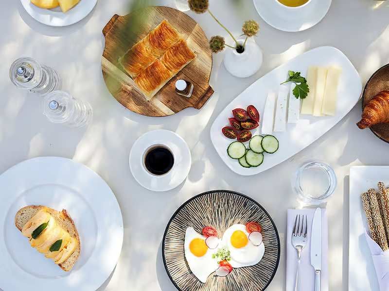 Gourmet breakfast spread including croissants, eggs, toast, cheese, cucumbers, cherry tomatoes, and coffee, arranged on a white table.
