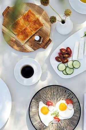 Gourmet breakfast spread including croissants, eggs, toast, cheese, cucumbers, cherry tomatoes, and coffee, arranged on a white table.