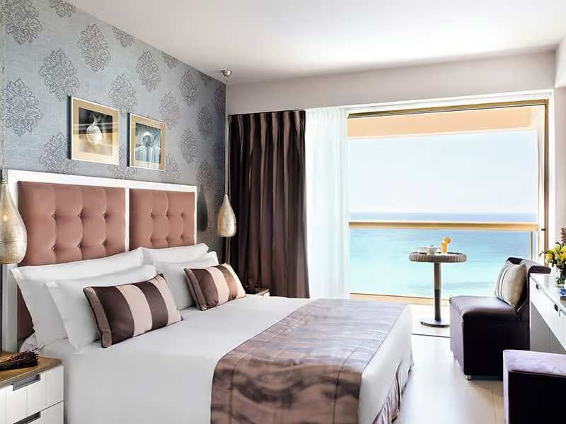 Luxurious hotel room with a double bed, modern decor, and a large window offering a view of the beach.