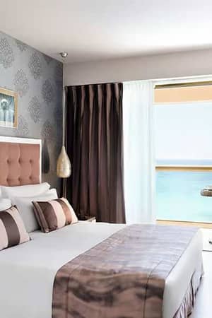 Luxurious hotel room with a double bed, modern decor, and a large window offering a view of the beach.