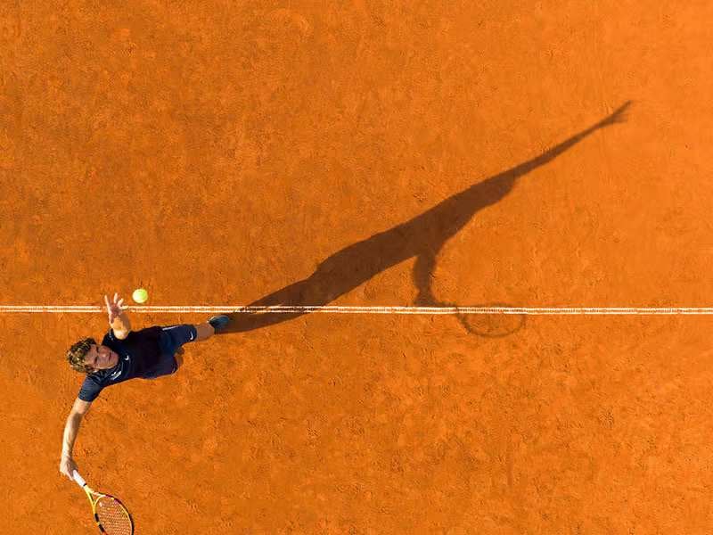 Overhead drone view of a tennis player serving on a clay court with a long shadow cast on the orange surface.