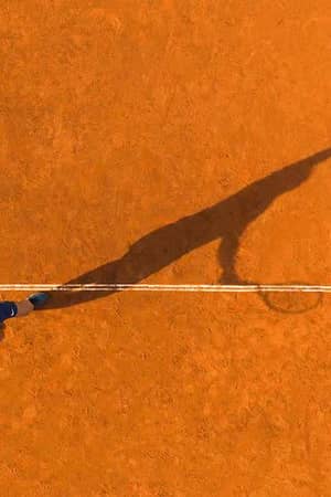 Overhead drone view of a tennis player serving on a clay court with a long shadow cast on the orange surface.