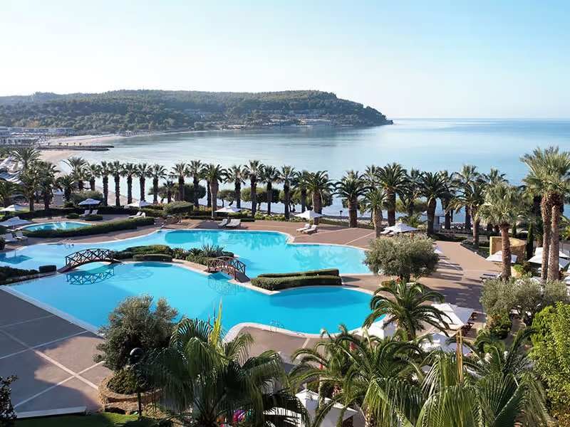 Wide shot of multiple swimming pools at Sani Beach School, surrounded by palm trees and overlooking a sandy beach and ocean.