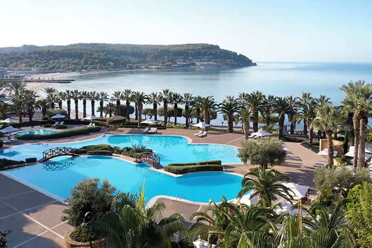 Wide shot of multiple swimming pools at Sani Beach School, surrounded by palm trees and overlooking a sandy beach and ocean.