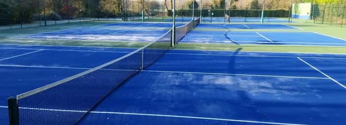 Tennis courts with a light frost covering, surrounded by bare trees, on a clear winter morning.