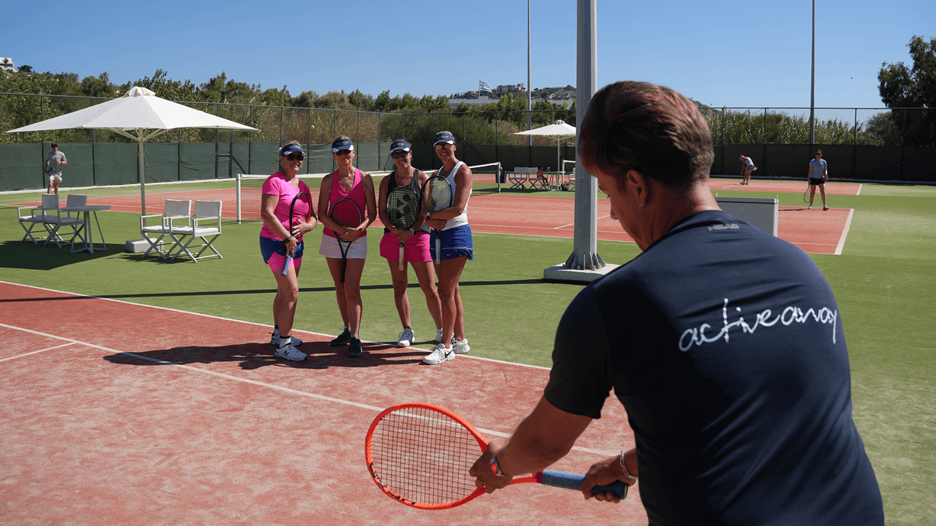 Tennis coach demonstrates to four players on a sunny outdoor court during an Active Away group lesson.