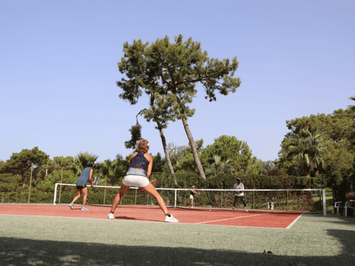 Four players in a doubles tennis game on an outdoor court surrounded by trees.
