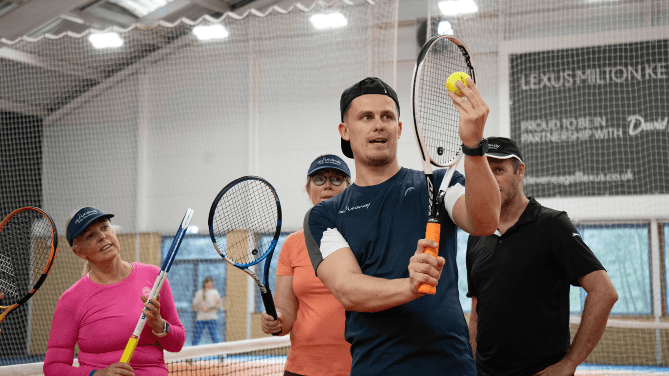 Coach demonstrating tennis shot with ball and racquet to adults during an indoor clinic.