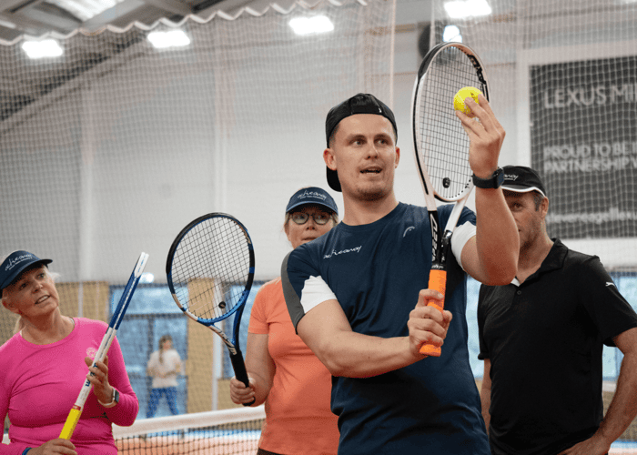 Coach demonstrating tennis shot with ball and racquet to adults during an indoor clinic.