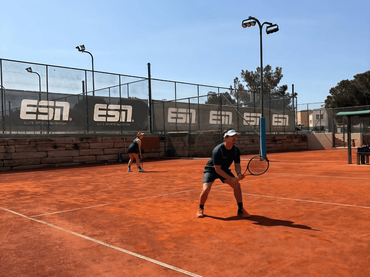 Two tennis players on a clay court in ready stance during a doubles match on a sunny day.
