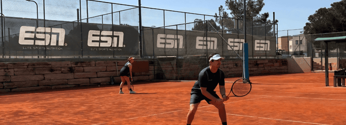 Two tennis players on a clay court in ready stance during a doubles match on a sunny day.