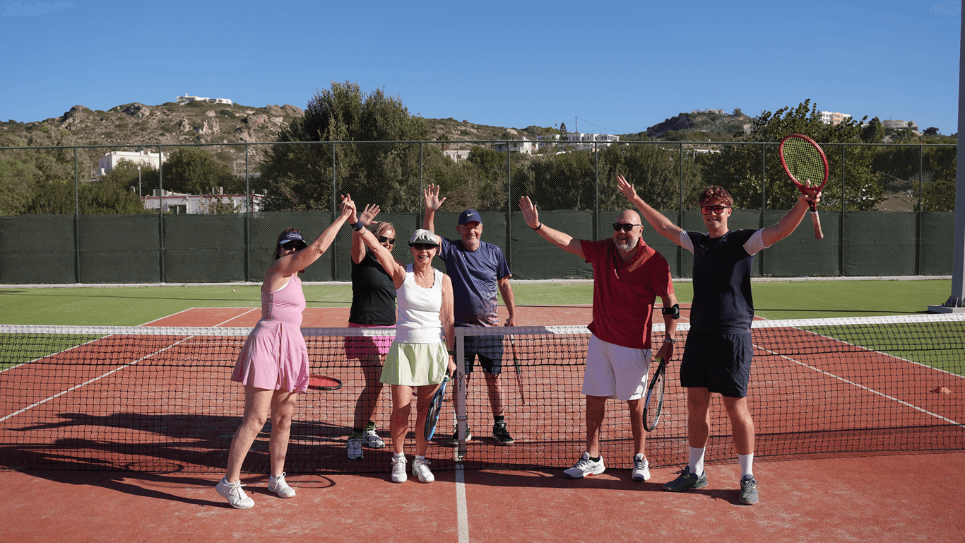 Six adults on a sunny tennis court raise their arms by the net, holding rackets and smiling.