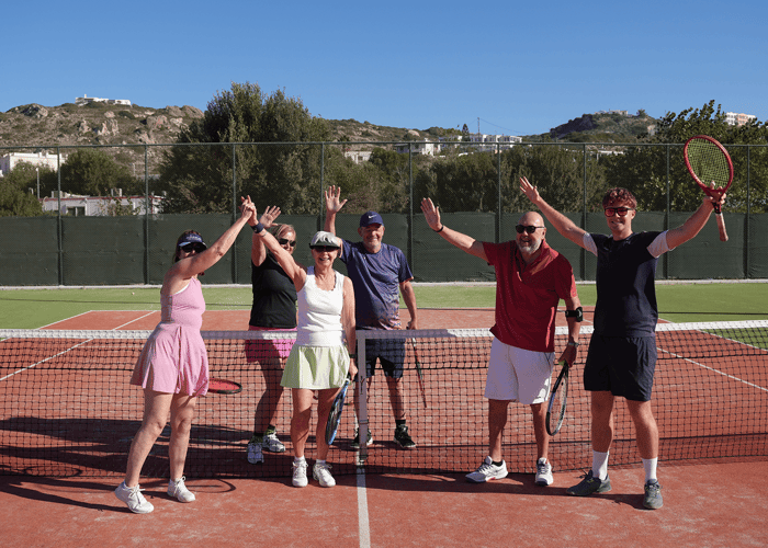 Six adults on a sunny tennis court raise their arms by the net, holding rackets and smiling.