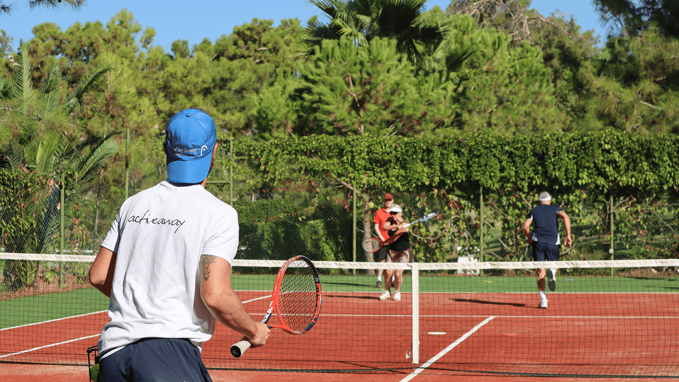 Coach observes doubles drill on a red hard tennis court as players rally near the net outdoors.