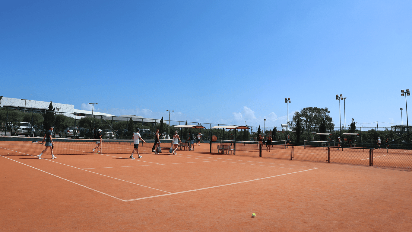Group tennis session on clay courts under a clear blue sky at a holiday resort.