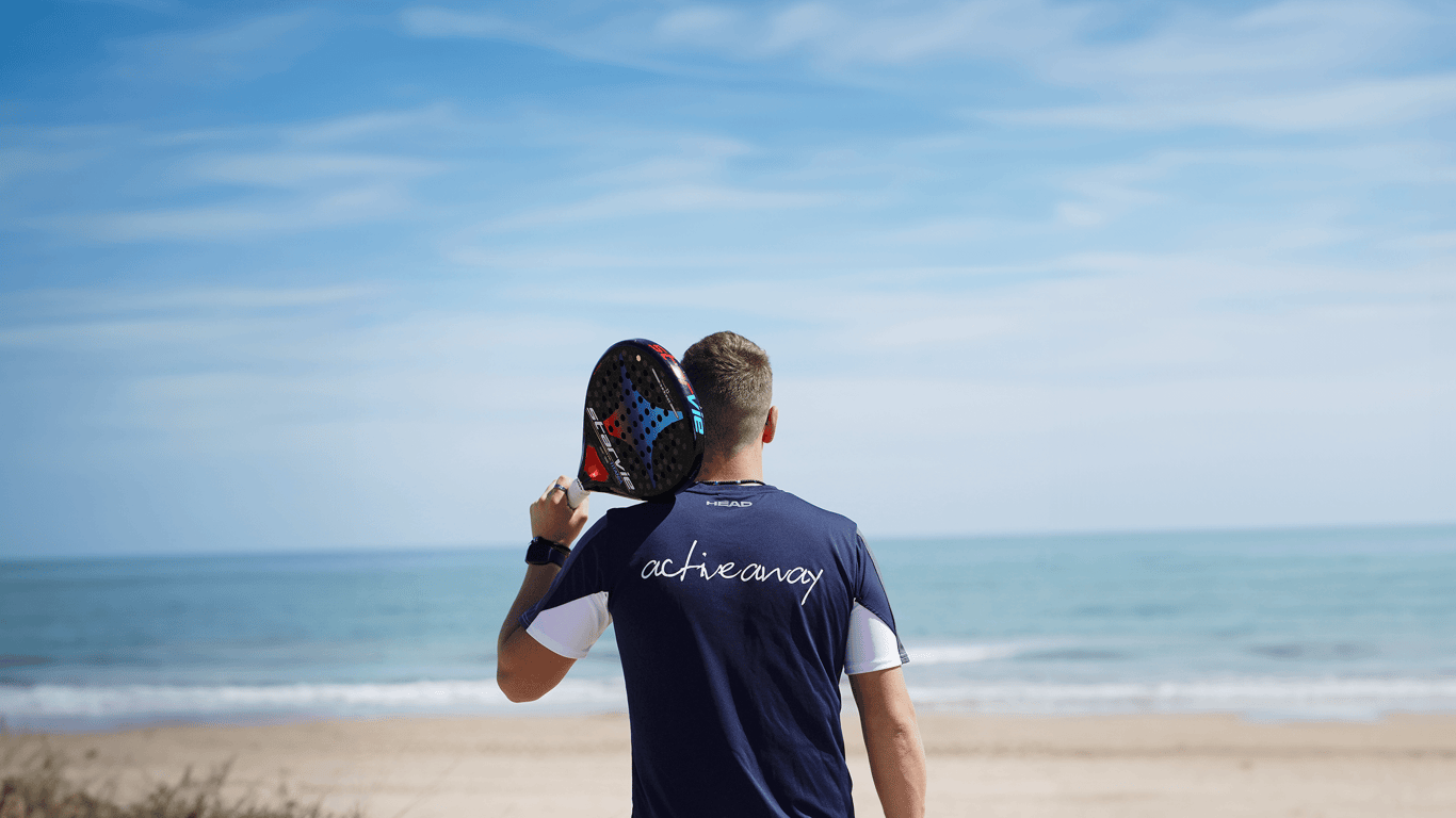 Man holding a padel racket on his shoulder, facing the sea, wearing an Active Away shirt.