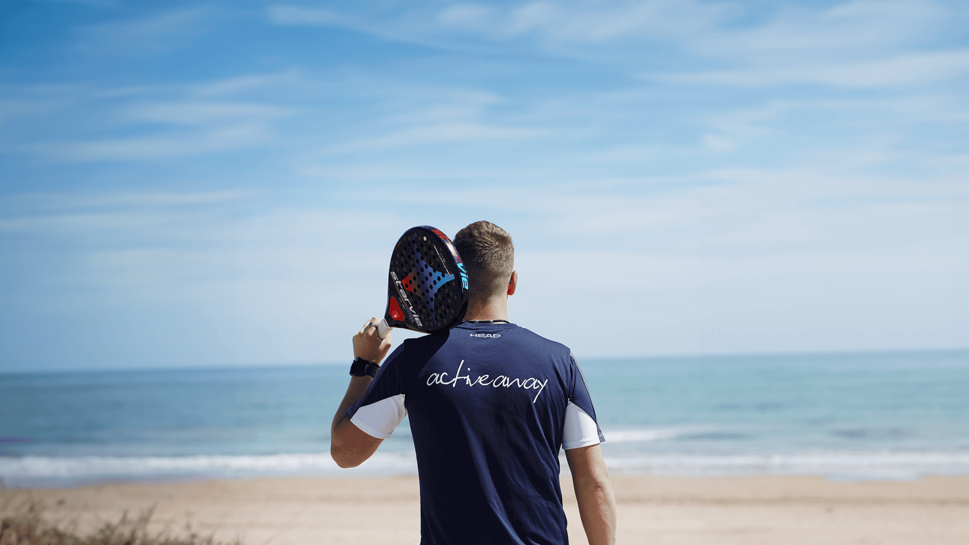 Man holding a padel racket on his shoulder, facing the sea, wearing an Active Away shirt.