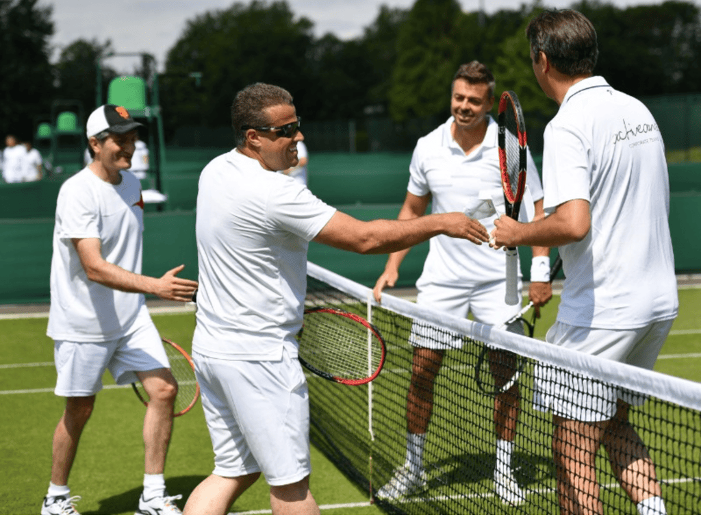 Four men in white tennis gear shake hands across a net on a sunny grass court.