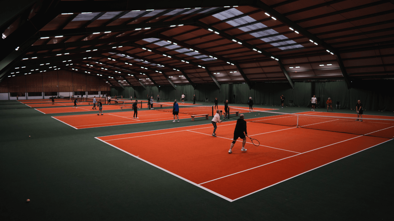 Busy indoor tennis centre with multiple orange courts and players practising drills under bright lights.