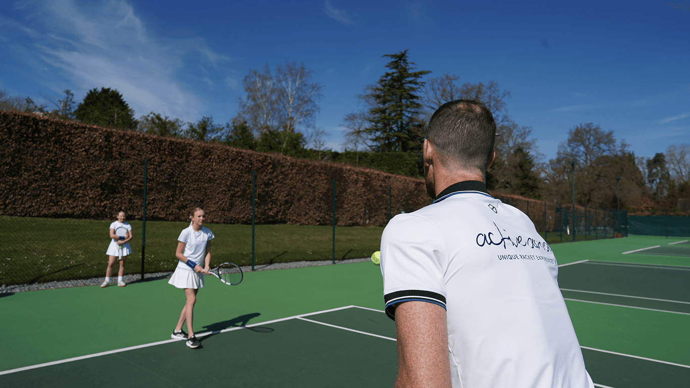 Tennis coach feeds a ball to a junior player during an outdoor lesson as another child waits to practise.
