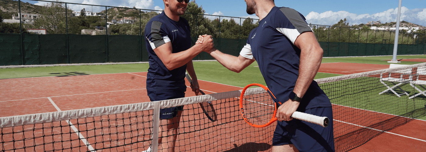 Two tennis coaches shake hands over the net on a sunny outdoor court.