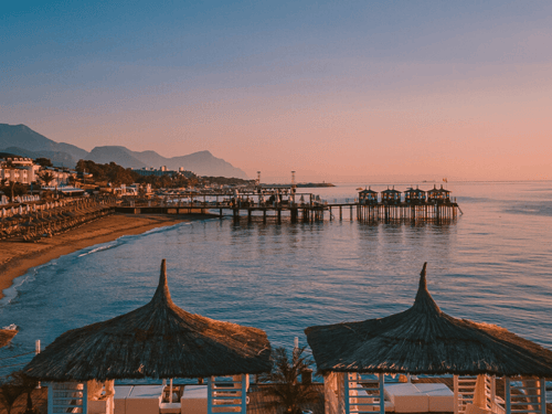 Sunset over a calm Mediterranean beach resort with jetties and cabanas, mountains on the horizon.