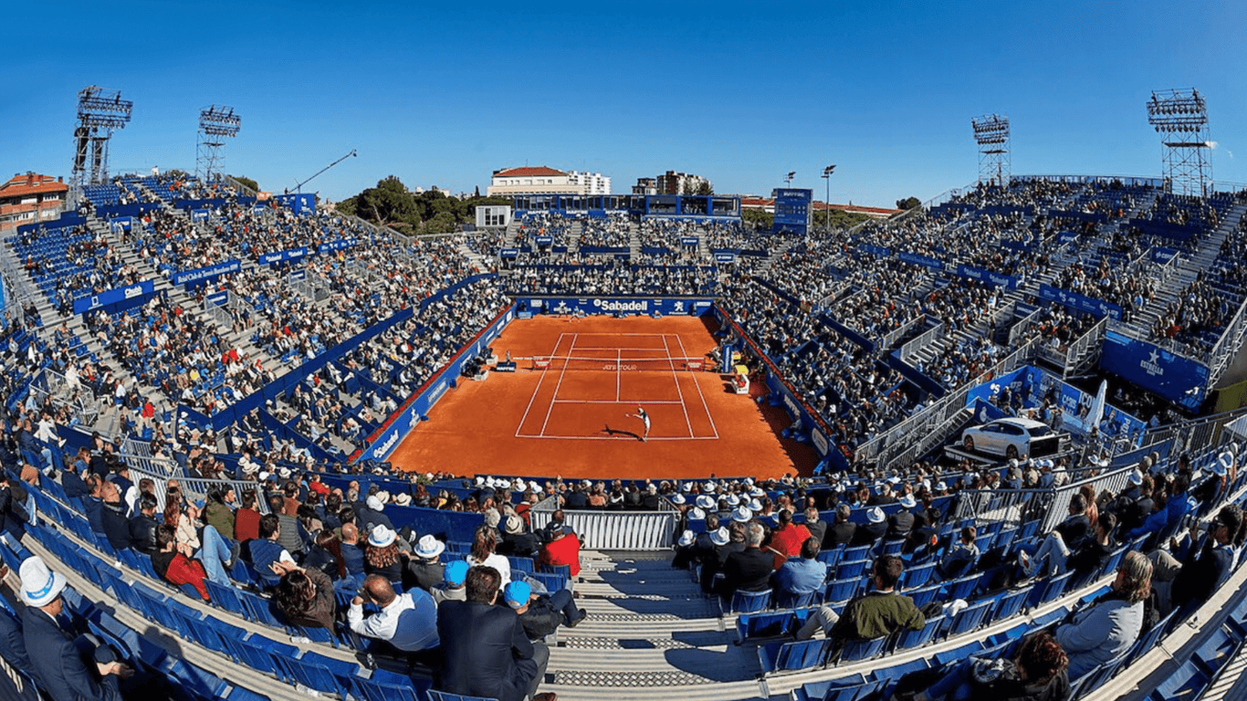 Aerial view of a clay-court tennis match at the Barcelona Open with packed blue stands and clear skies.