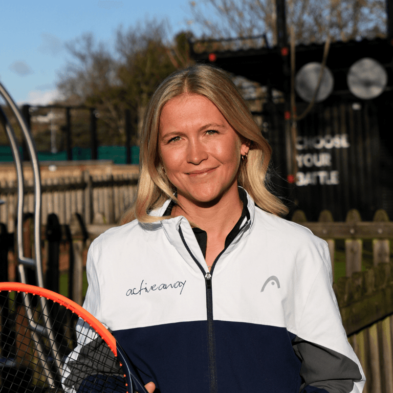 Smiling tennis coach holding a racket outdoors in an Active Away jacket.