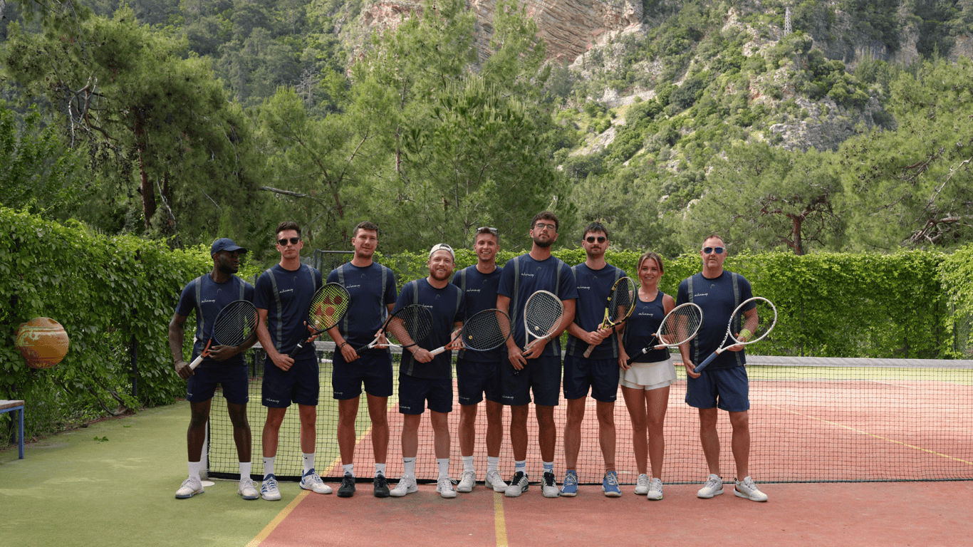Line of Active Away tennis coaches holding rackets on an outdoor court with forested mountains behind.