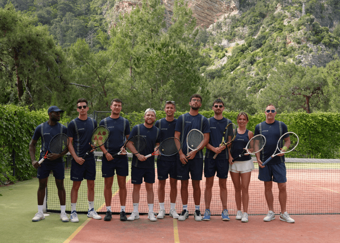 Line of Active Away tennis coaches holding rackets on an outdoor court with forested mountains behind.