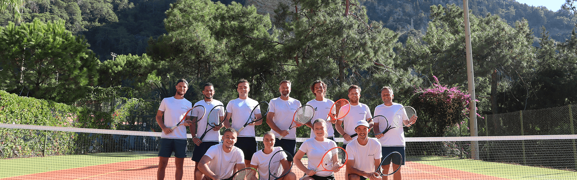 Group of tennis players posing with rackets on a sunny court, mountains and pine trees in the background.