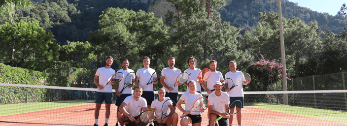 Group of tennis players posing with rackets on a sunny court, mountains and pine trees in the background.