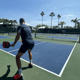 Player prepares a shot on an outdoor pickleball court under blue sky and palm trees.