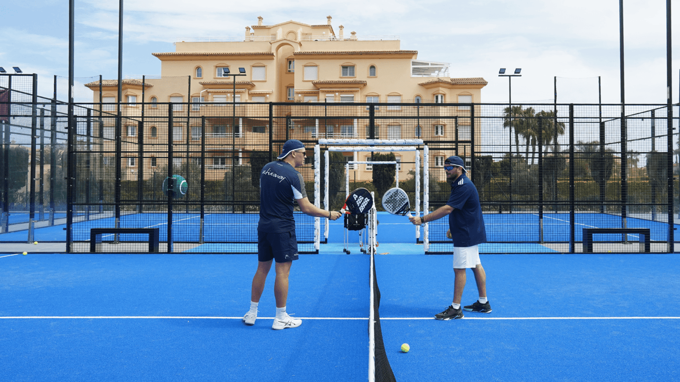 Two padel coaches bump rackets across the net on a blue Oliva Nova court.