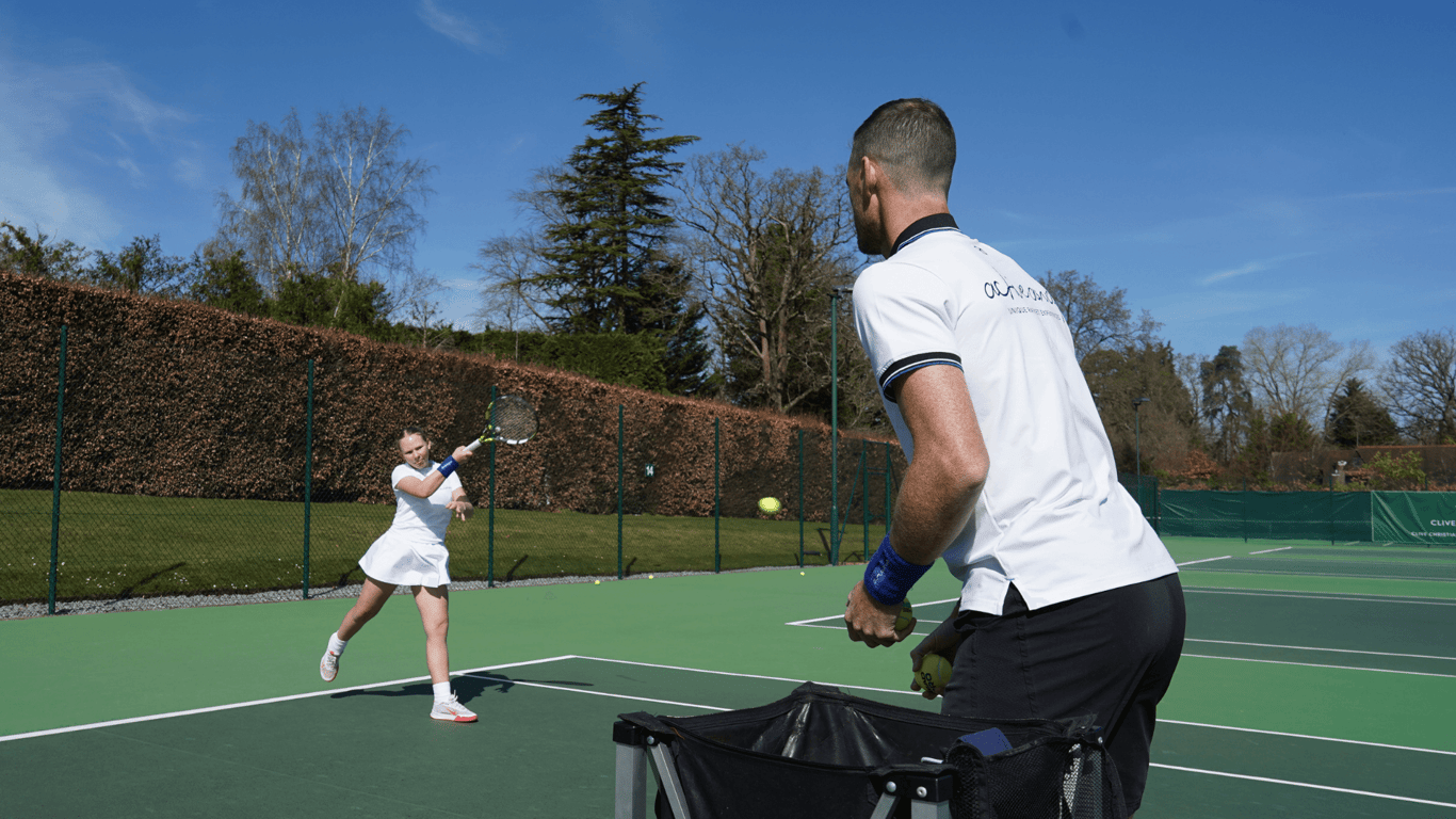 Coach feeds balls from a basket as a young player practises a forehand on an outdoor tennis court.