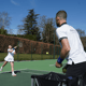 Coach feeds balls from a basket as a young player practises a forehand on an outdoor tennis court.