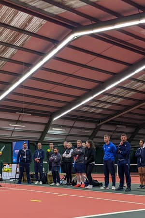 Indoor tennis training at David Lloyd West Bridgford with players watching a coach demonstrate a drill.