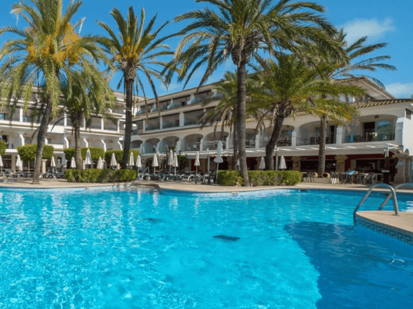 Sunlit resort pool with palm trees, loungers and a white hotel building in the background.
