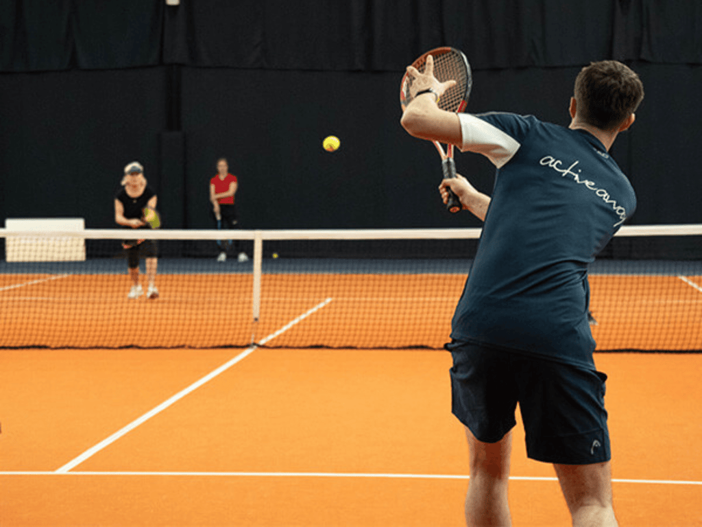 Indoor tennis session: player prepares a backhand volley as opponents wait across the net.