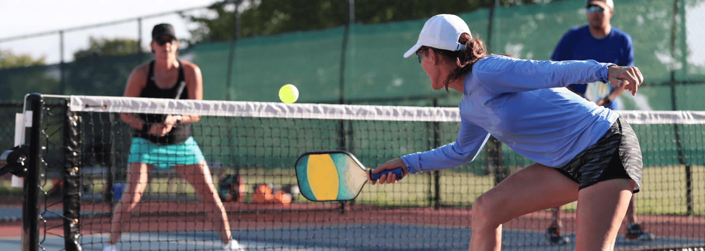 Woman lunges to hit a pickleball at the net during a doubles game on an outdoor court.