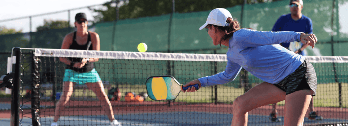 Woman lunges to hit a pickleball at the net during a doubles game on an outdoor court.