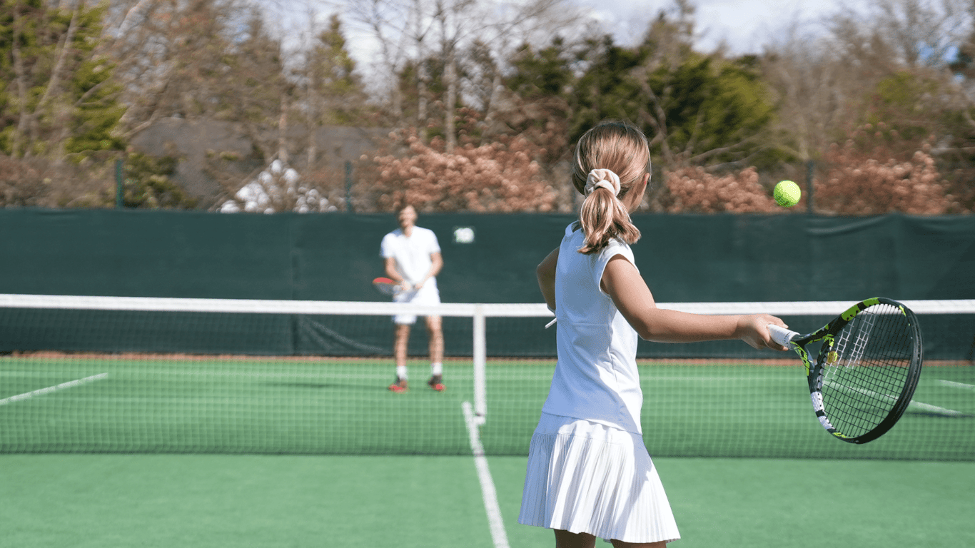 Young girl practising a forehand during a tennis lesson on an outdoor court with a coach across the net.