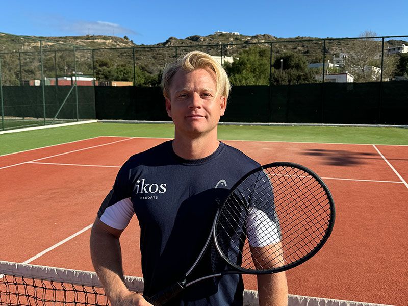 Tennis coach holding a racket on an outdoor court at Ikos Aria resort.