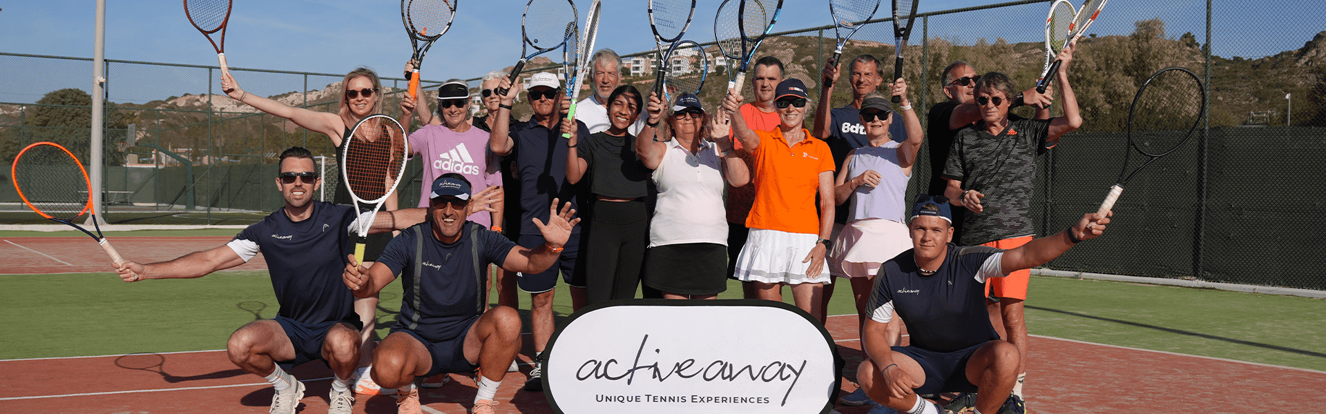 Group of tennis players on court raising rackets, posing with Active Away sign under sunny sky.